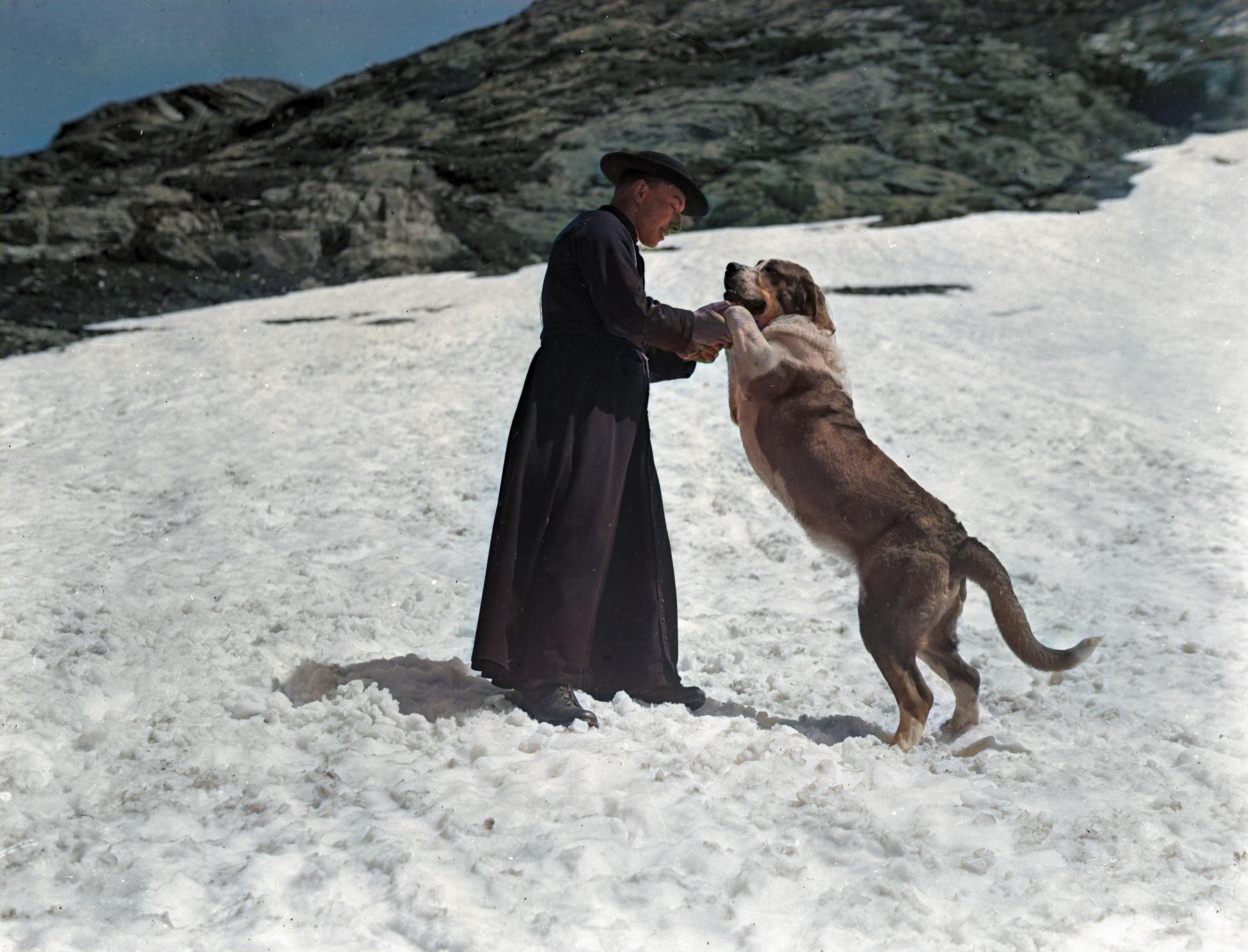 A monk in a dark robe stands on a snowy slope, holding the front paws of a large St Bernard dog that is rearing playfully on its hind legs.