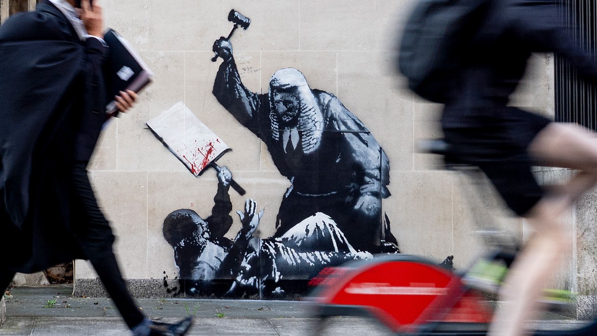 A photograph captures a person in a judge's robe walking while on a phone call, passing by a Banksy mural on a building wall that depicts an old, robed man wielding a hammer over a child holding a book.