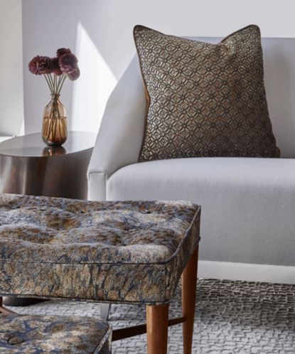 Close up of cream sofa with brown patterned cushion, textured cream rug, footstool with wooden frame and brown upholstery, metal side table with vase in background