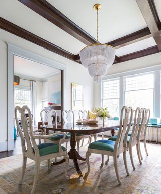 Light and bright dining room with white painted walls, dark wood ceiling beams, dark wood flooring, large, traditional patterned rug, traditional glass chandelier, large dark wood dining table with light wood dining chairs