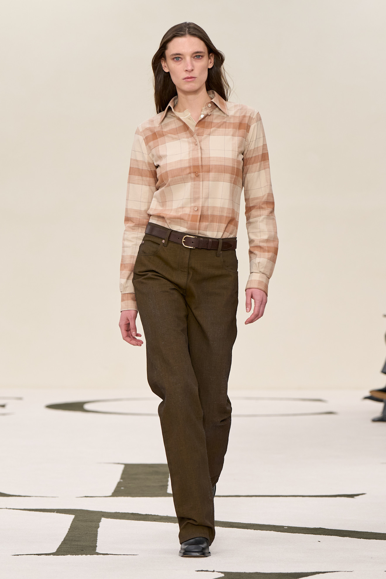 A look from Calvin Klein's F/W 25 runway show including a tan plaid shirt tucked into brown-green pants.