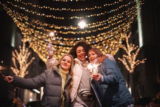 A group of three friends holding sparklers in front of Christmas lights.