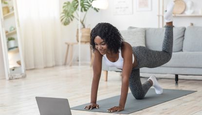 Woman exercising at home. She is on her hands on an exercise mat, with one leg lifted behind her. She is looking at a laptop in front of her and is wearing a white top, grey leggings and white socks.