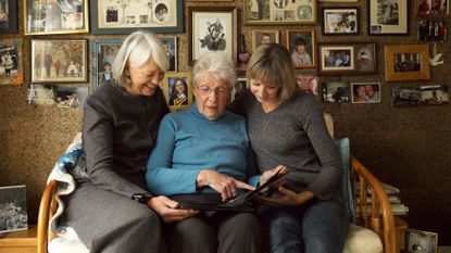 Three women of different ages sit together on a couch and point to a photo in a photo album