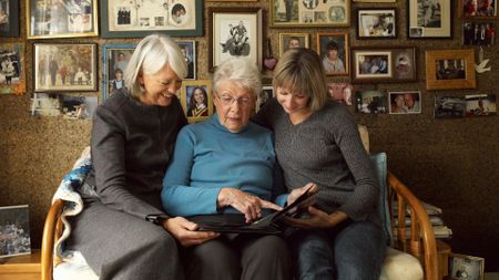 Three women of different ages sit together on a couch and point to a photo in a photo album