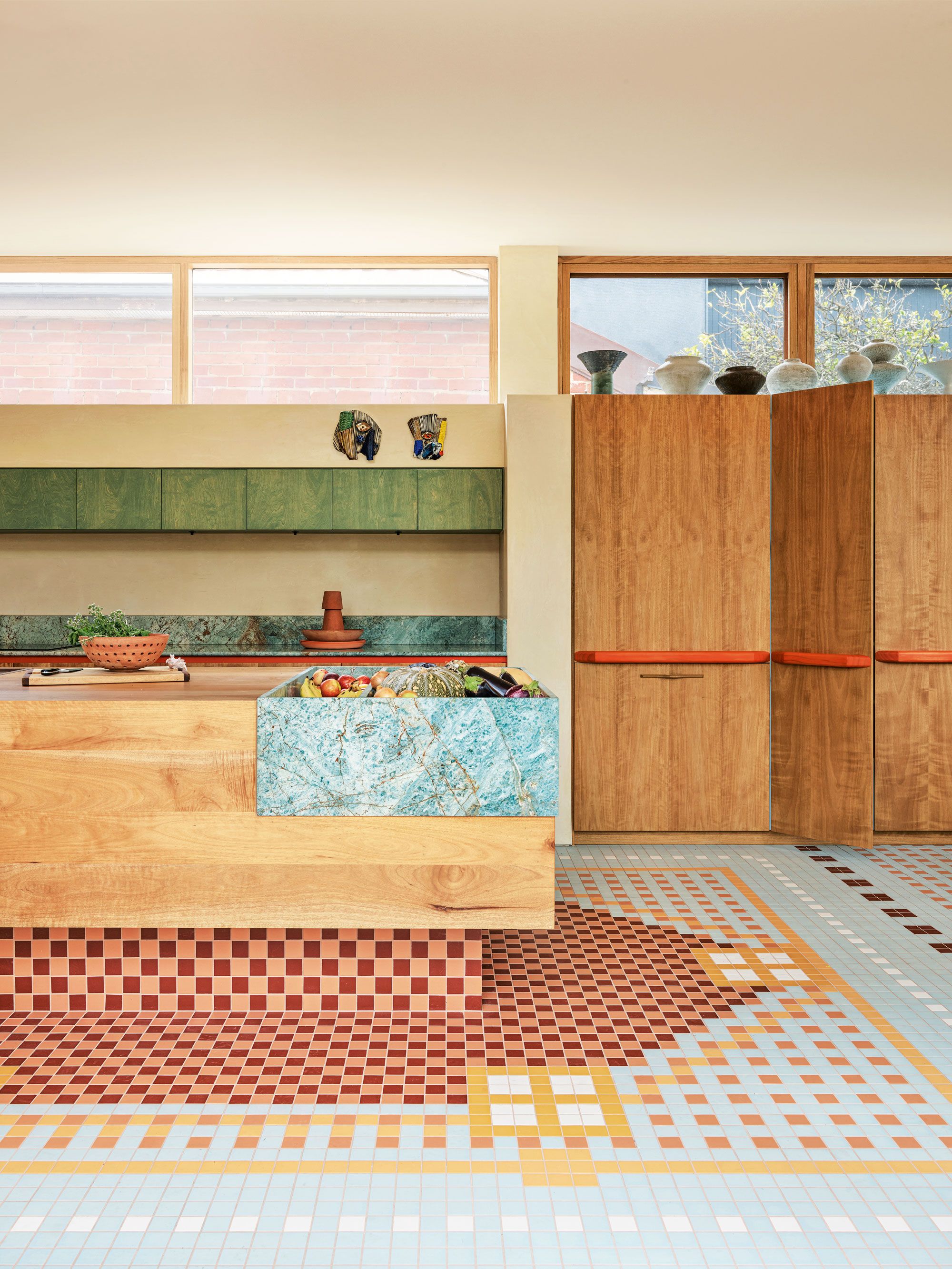 Bright, open kitchen are with high windows and wood cupboards and storage underneath. A wooden islands stand in the middle of the space with a custom turquoise, marble feature. Below, a geometric, rug style tile flooring in bright blue and earthy tones. 