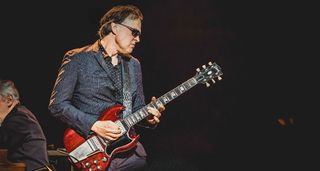 Joe Bonamassa plays a vintage Gibson Les Paul SG equipped with a Maestro Vibrola. He wears a gray patterned suit and sunglasses.