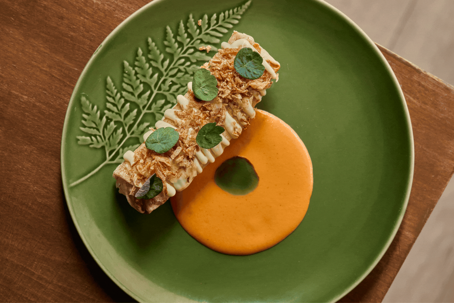 A green ceramic plate filled with a beautiful vegetarian dish composed of an orange mousse, green oil, and a crunchy onion-topped mousse sits atop a wooden table.