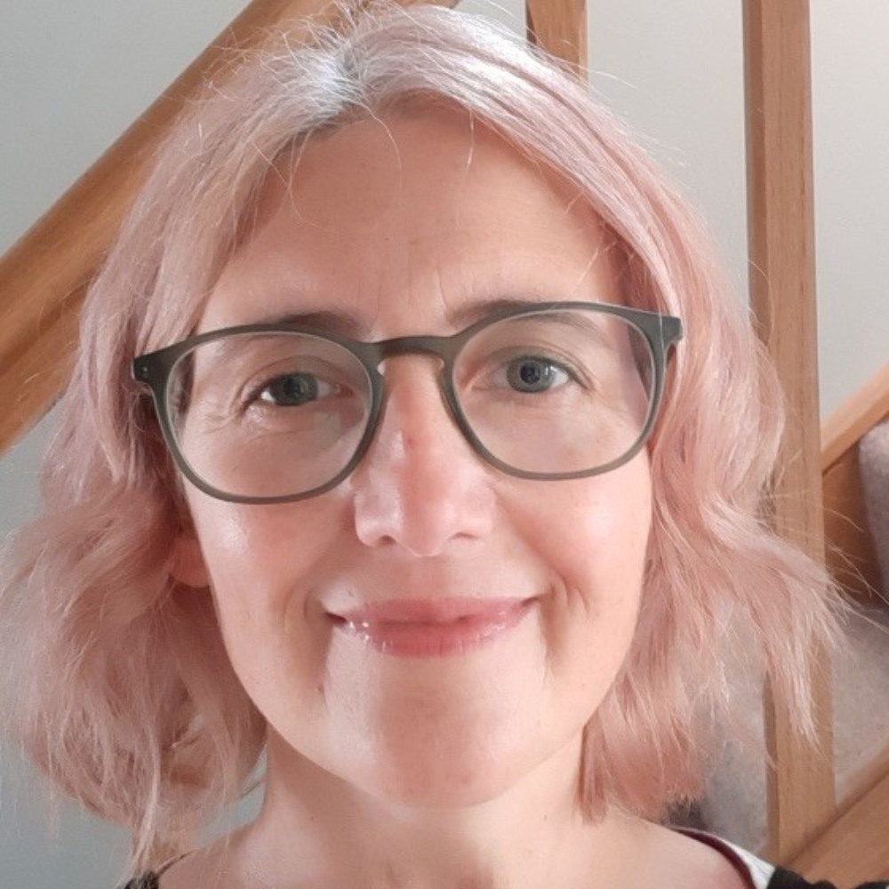 Smiling woman with pink-tinted hair and round glasses, close-up headshot against a wooden staircase backdrop.