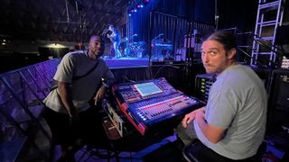 Two sound engineers sit at a DiGiCo mixing console showered in purple and blue lights at a concert.