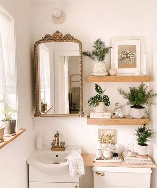 A white bathroom with wooden shelves with wall art and plants on it, a vintage arched mirror, a sink with brass faucets, and a wooden shelf with candles on top of it