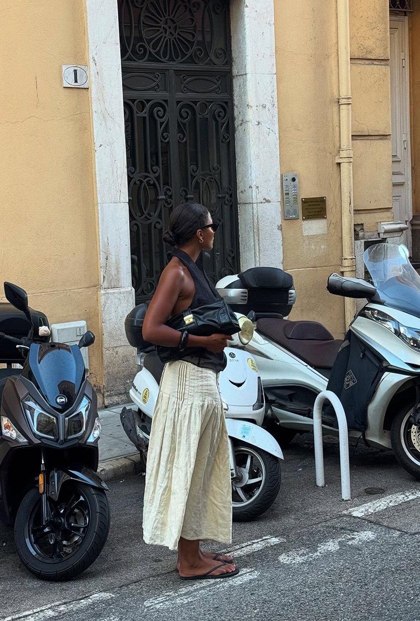 A photo of a woman standing outside wearing a black halter top with a tan maxi skirt, black flip-flops, black clutch bag, and black sunglasses