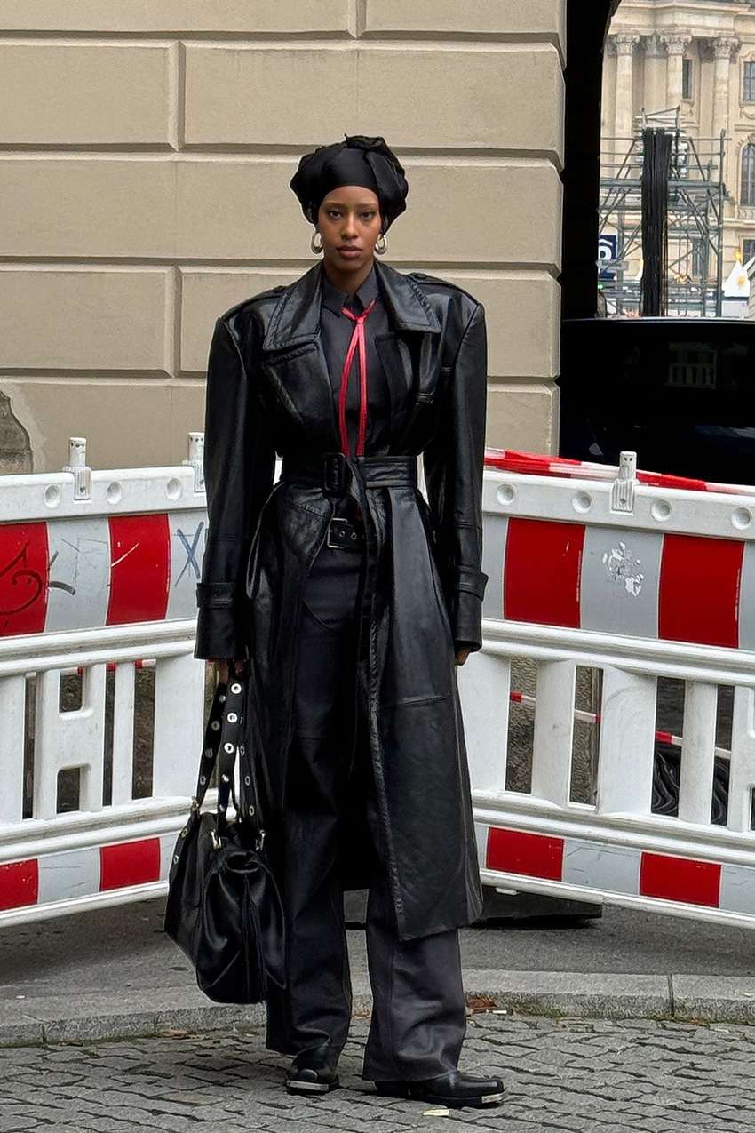 Berlin fashion trends are shown in a street style photo of a woman standing in the street wearing a black headscarf, hoop earrings, a long black trench coat over a gray collared shirt styled with a thin red scarf, black leather pants, black square toe ankle boots, and a black studded handbag