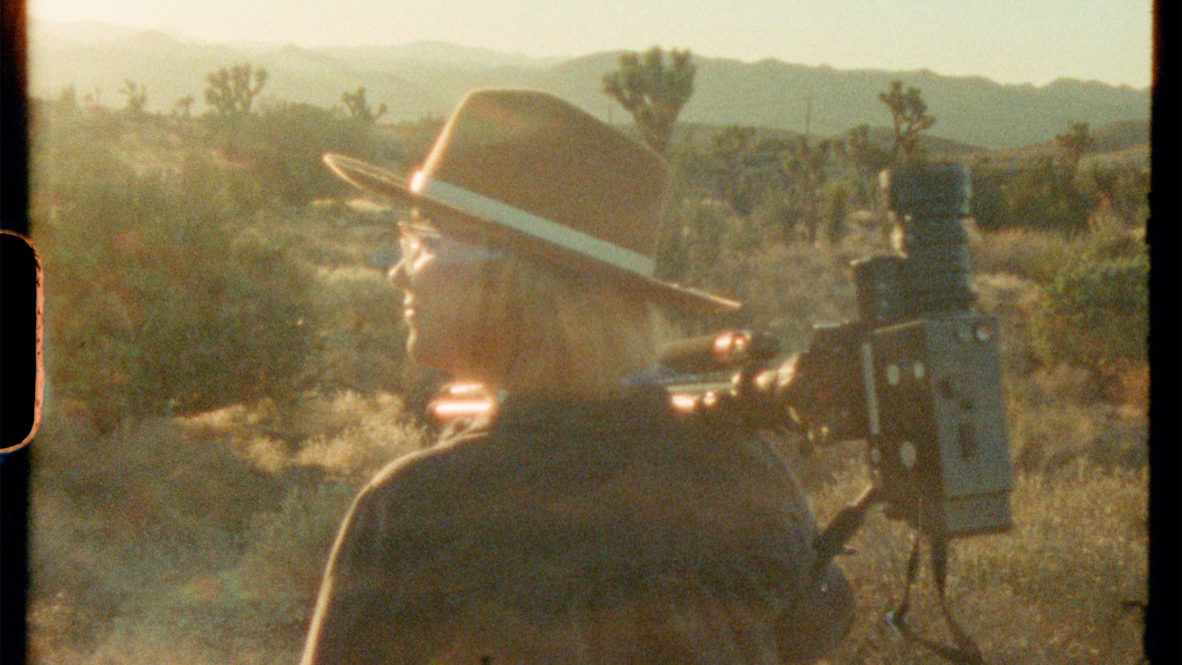 A man looks toward the sun while holding a film camera on a tripod over her shoulder.