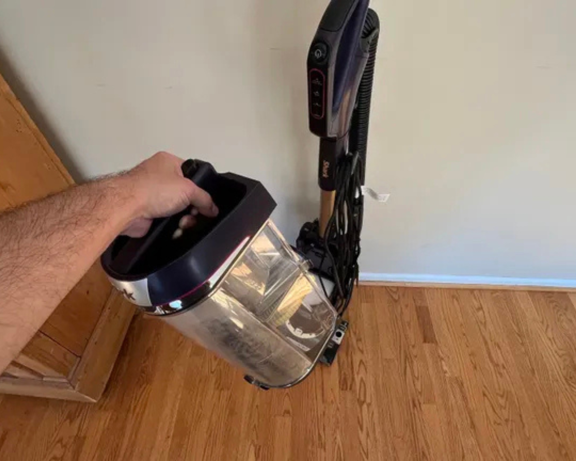 Hand holding the Shark POWERDETECT Upright Vacuum's dust bin in front of the vacuum on wooden floor.