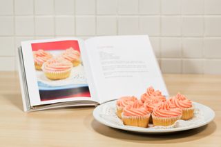 A plate of pink cupcakes with a recipe book page of the same pink cupcakes open on a cookbook stand in a kitchen.