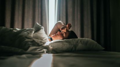 Woman lying on back in bed after night&#039;s sleep, hand on her forehead and sunlight coming through the window through the curtains