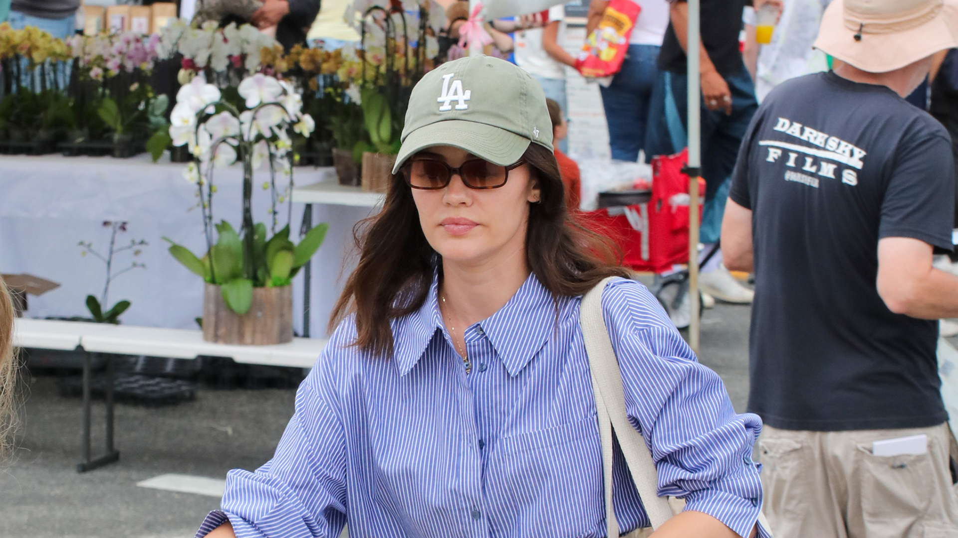 Lucy Hale wearing a white striped button down, gray jeans, nude sandals, and a tote bag, and a green LA cap