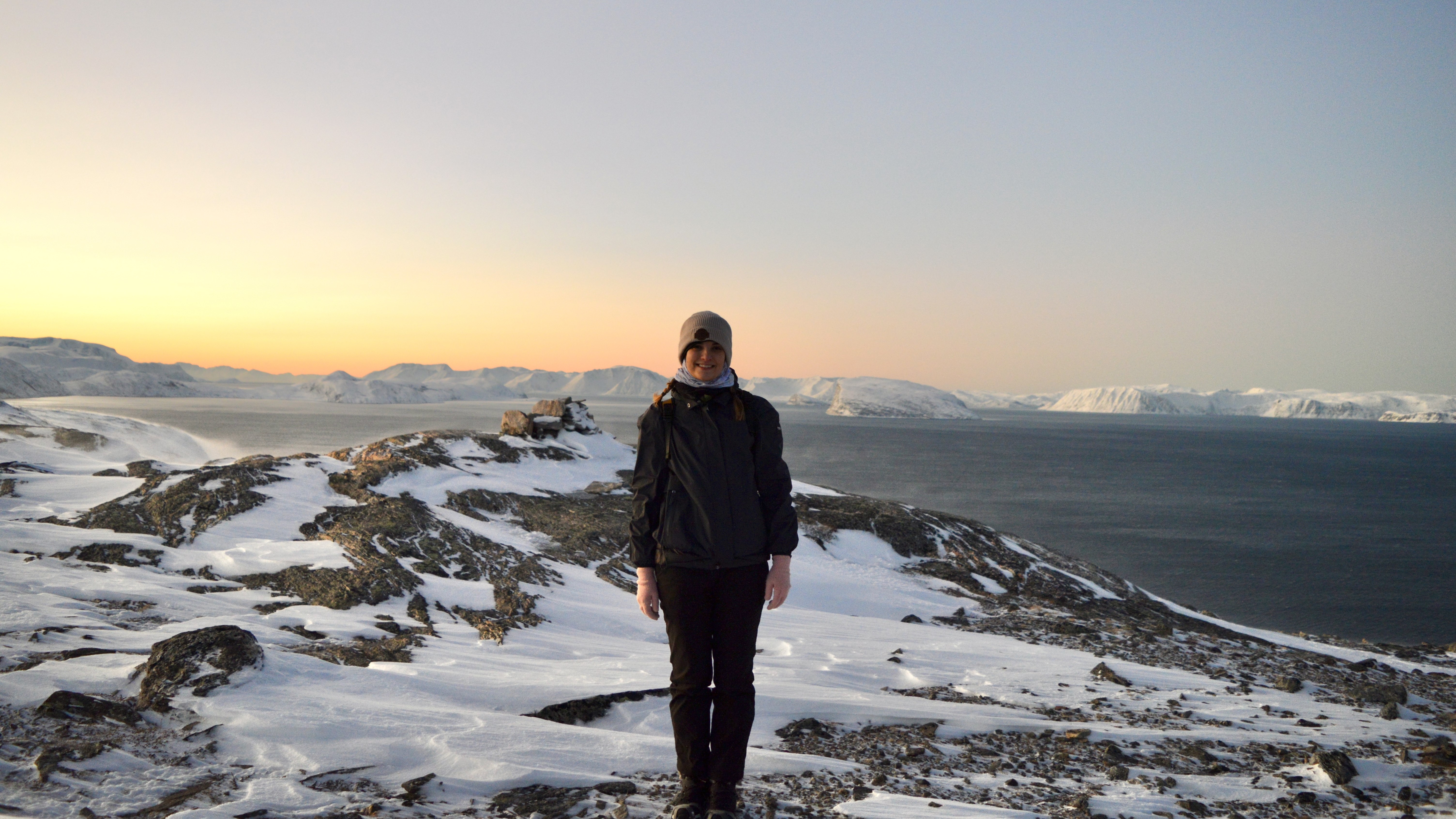 A woman is standing in the center of the image with a snowy mountain underfoot and the sea in the distance.