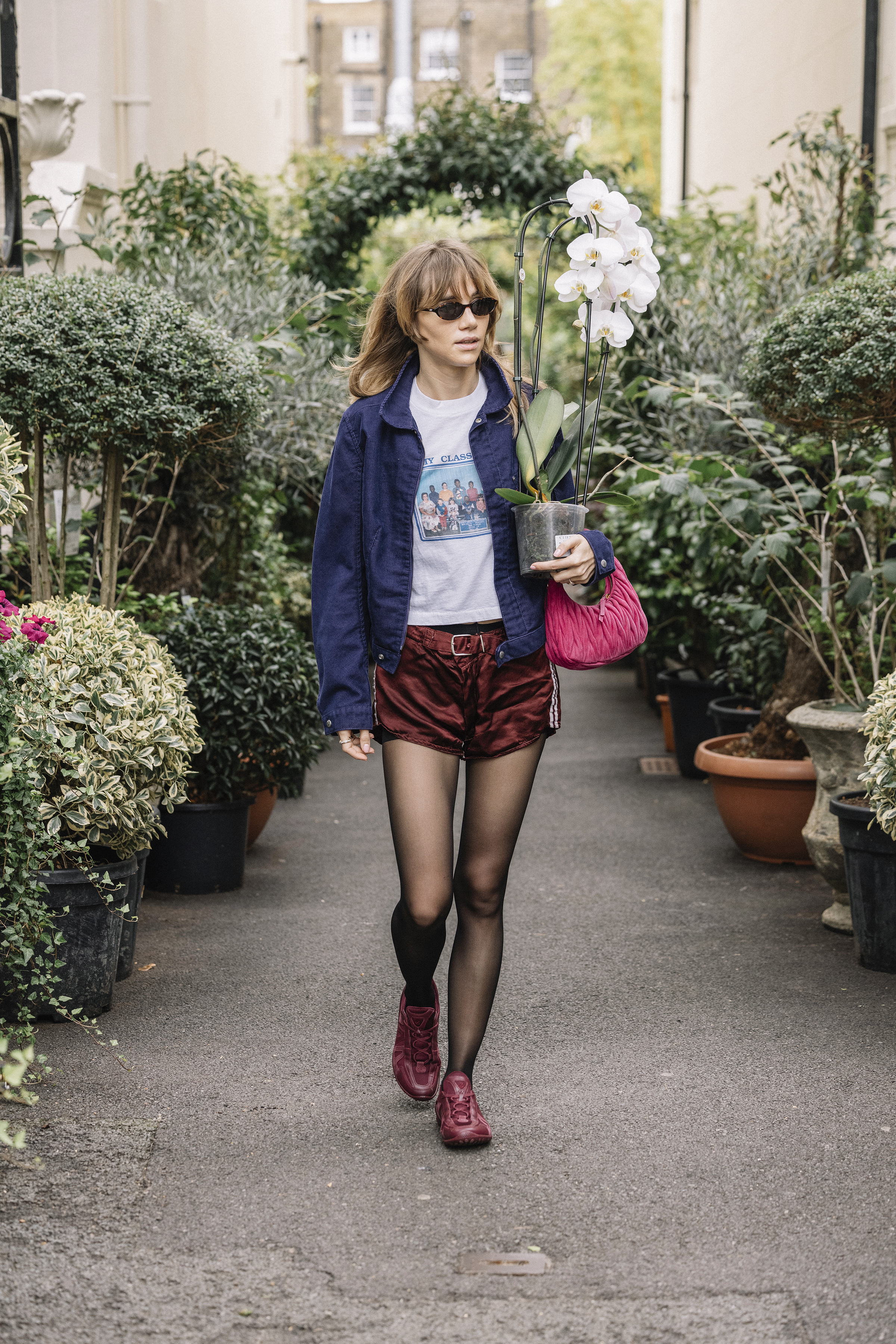 Suki Waterhouse viste una chaqueta azul marino, una camiseta blanca, pantalones cortos y zapatillas burdeos mientras lleva una planta en Londres.