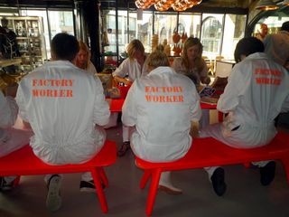 6 people sitting on an orange park bench in a restaurant wearing a white rob with the text Factory worker on the back