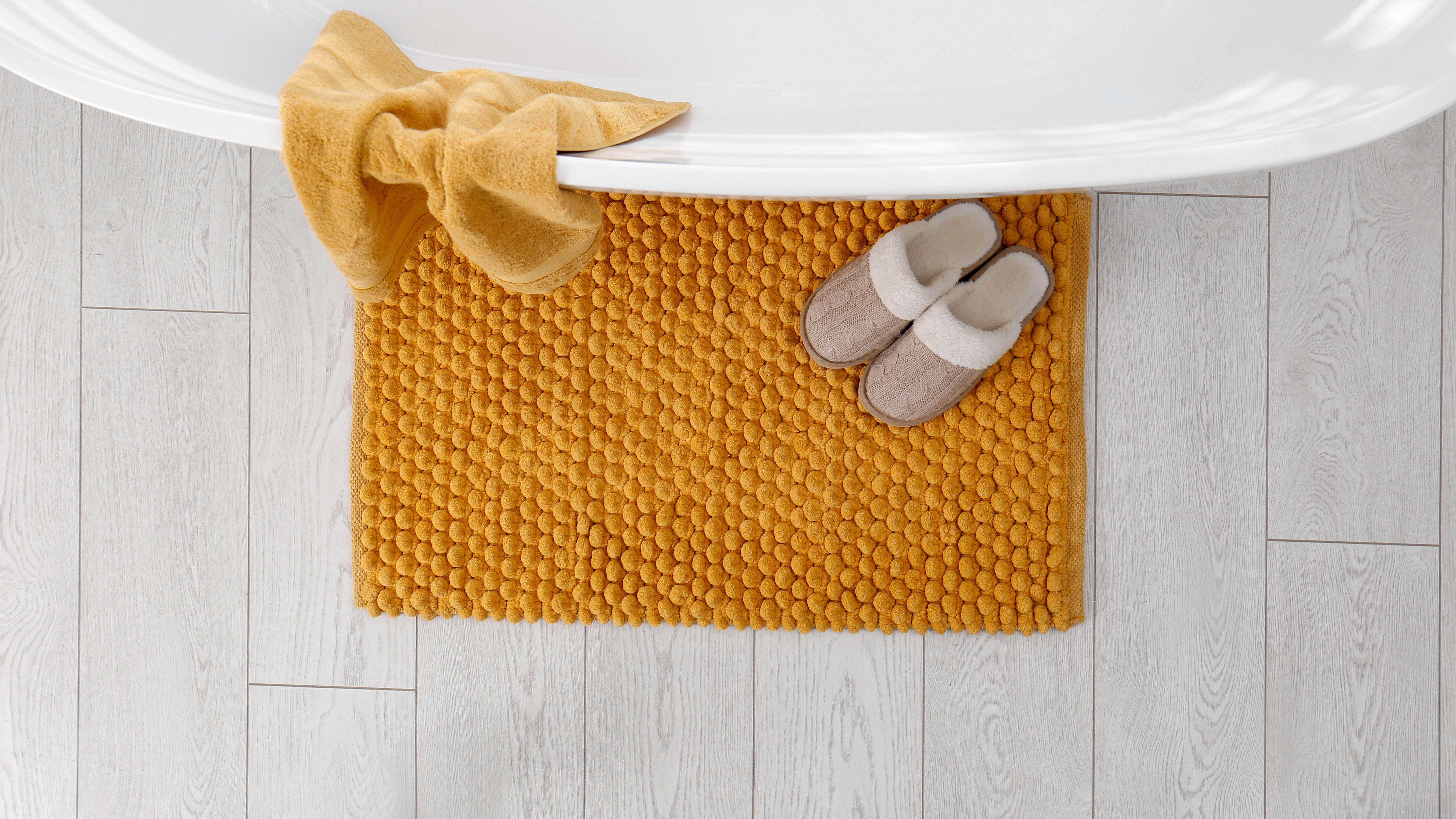 A yellow bath mat next to the bath tub with a pair of slippers and a towel