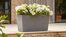 wheeled gray garden planter filled with flowers on a deck with stone detailing and brick wall behind
