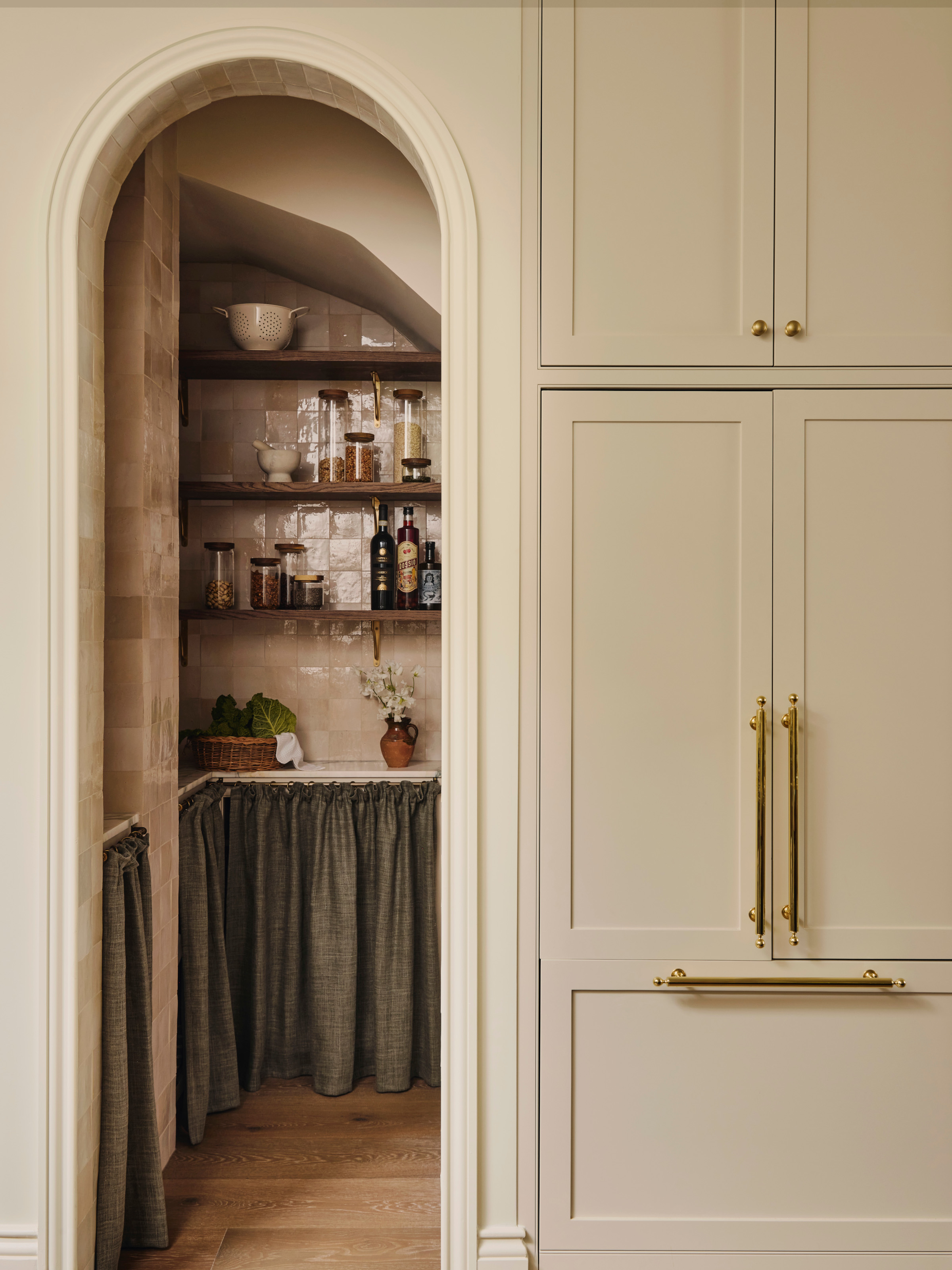 Pantry area with pale pink zellige tiles and dark sage green cupboard curtains tucked away behind an arched opening from a Shaker kitchen