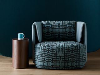 A blue armchair with a black pattern on it. Next to it is a cylinder-shaped wooden side table with a mug on it.