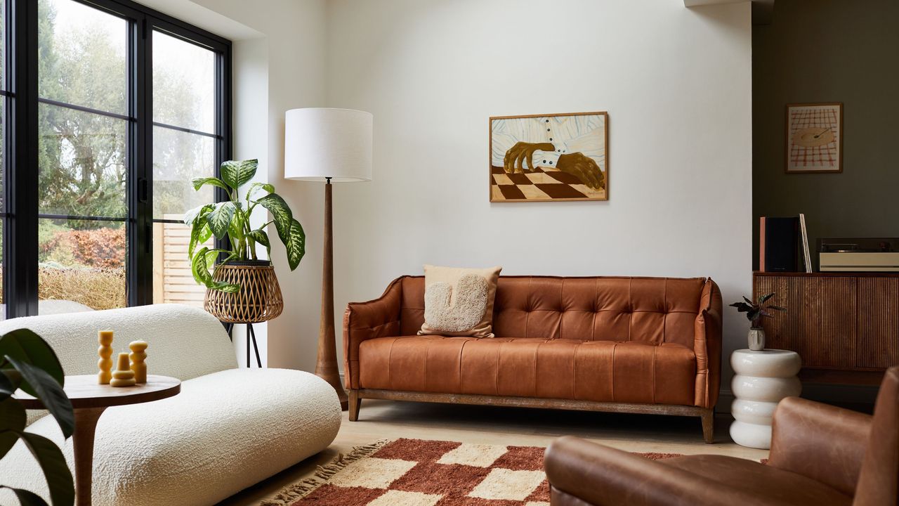 We&#039;ve created your checklist for moving into a first house. Here is a white living room with a tan couch with a brown wall art print above it, a tall white lamp, and a light and dark brown checked rug