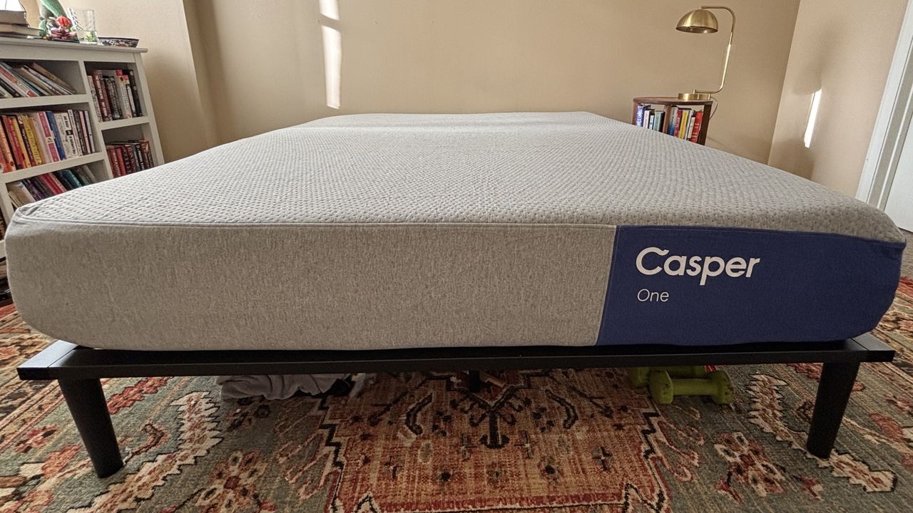 A queen-size Casper One Memory Foam Mattress, shown from the foot of the bed with the brand's purple and white logo on the right side. The mattress is on a black bed frame that's on top of an ornate bedroom rug with shades of red and green.