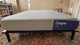 A queen-size Casper One Memory Foam Mattress, shown from the foot of the bed with the brand's purple and white logo on the right side. The mattress is on a black bed frame that's on top of an ornate bedroom rug with shades of red and green.