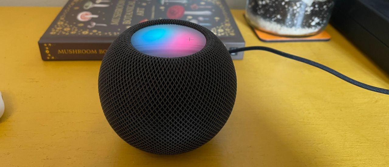 A dark grey Apple HomePod Mini on a piece of yellow furniture. The top panel of the speaker is illuminated in pink and blue colours.