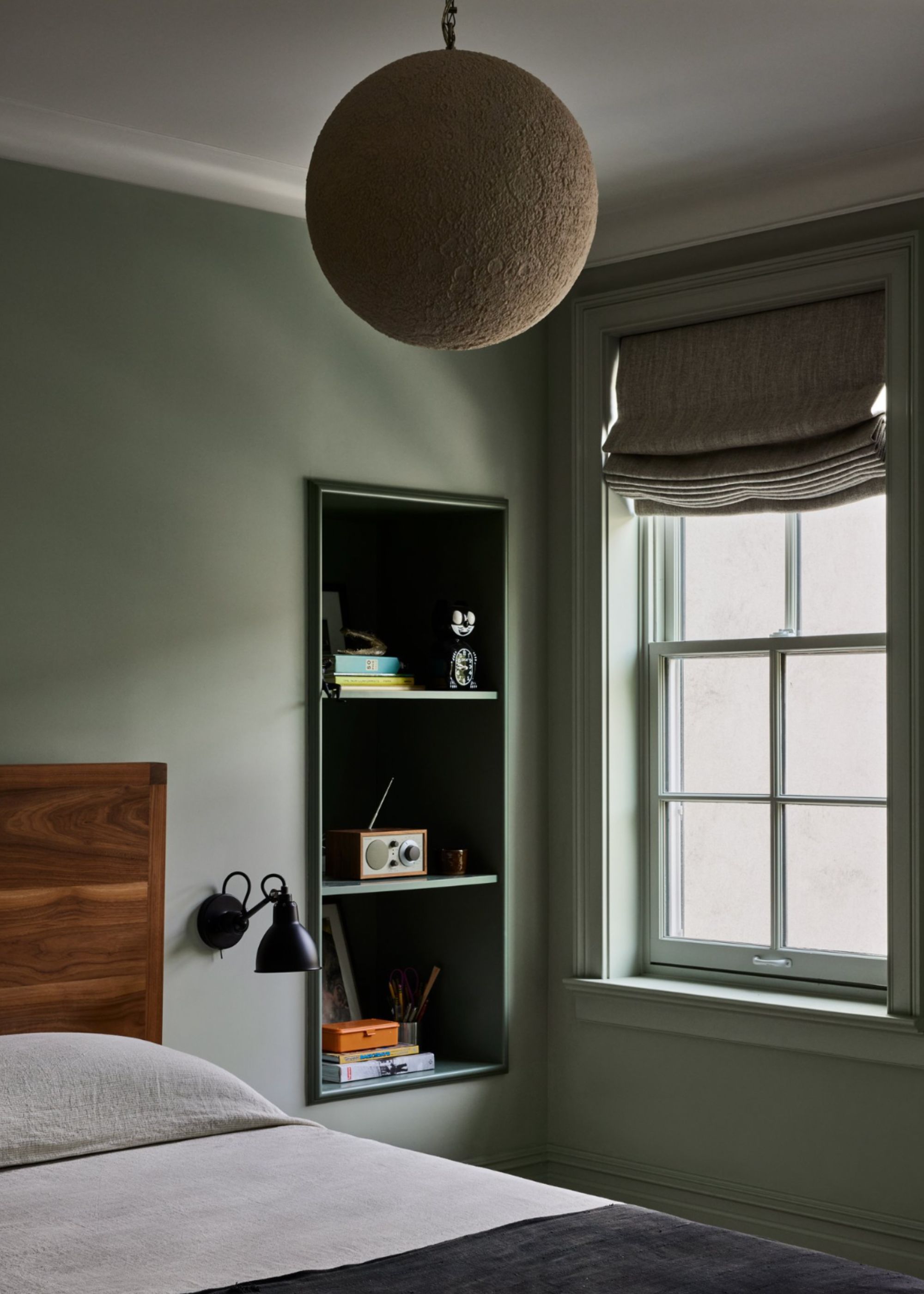 A bedroom with green wall paint, a round textured beige pendant light and roman blinds, alcove shelving and gray bedding
