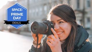 A smiling woman looking through the viewfinder on a Sony A7 III with buildings in the background. 