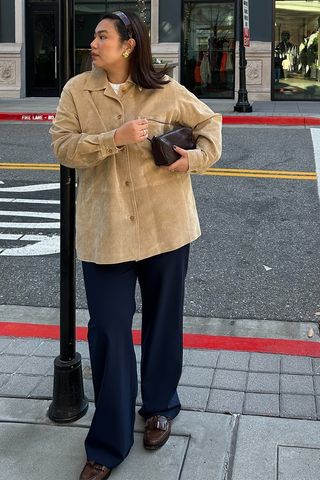 how to make a button-down shirt look more expensive is shown in a photo of a woman standing outside on the sidewalk wearing a brown headband, gold earrings, a brown suede button-down shirt over a white t-shirt styled with a brown barrel bag, dark-wash wide-leg denim, and dark brown loafers