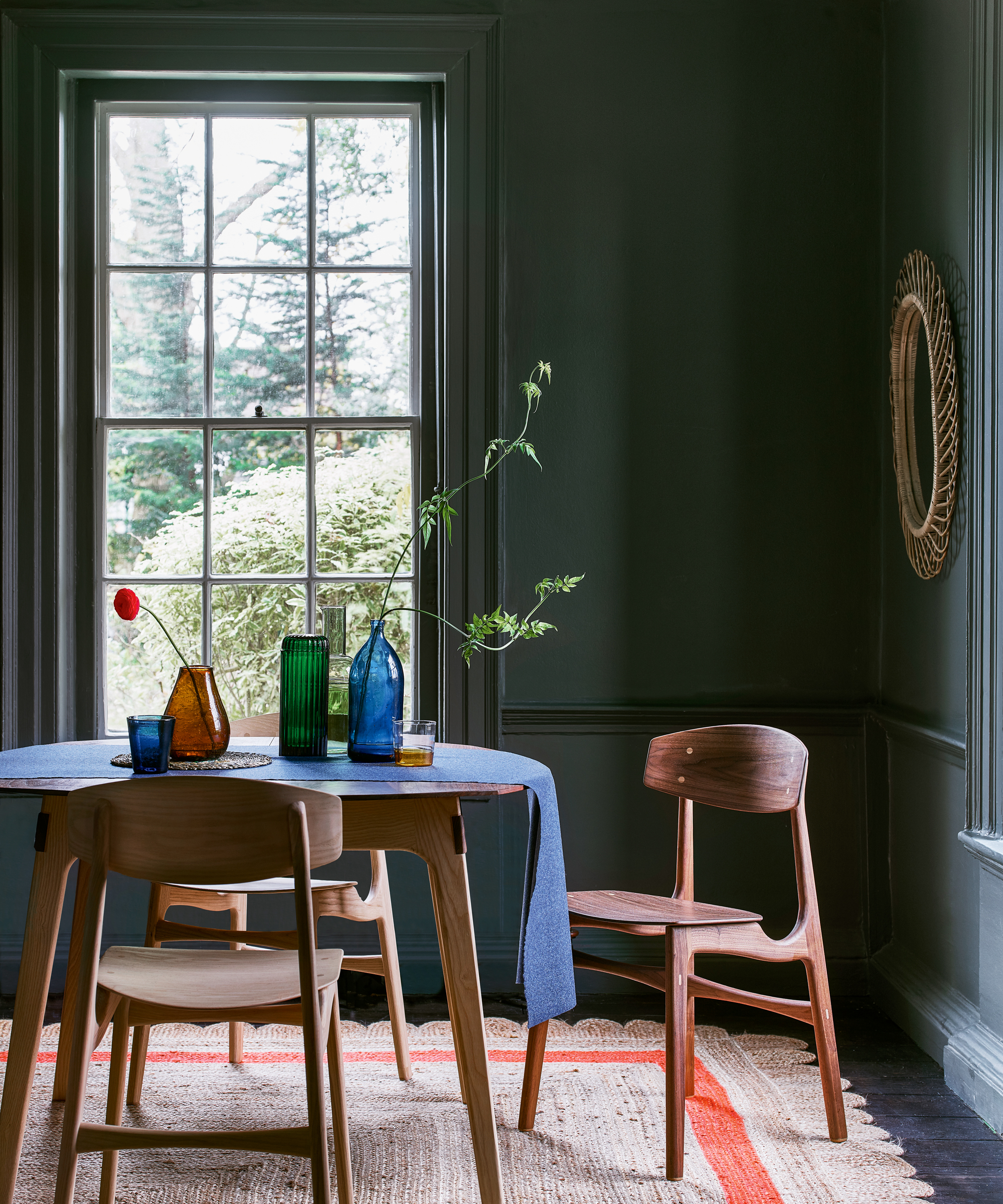 A dining room color idea with emerald green walls, rattan framed mirror, wooden chairs and table in a modern design and natural weave rug