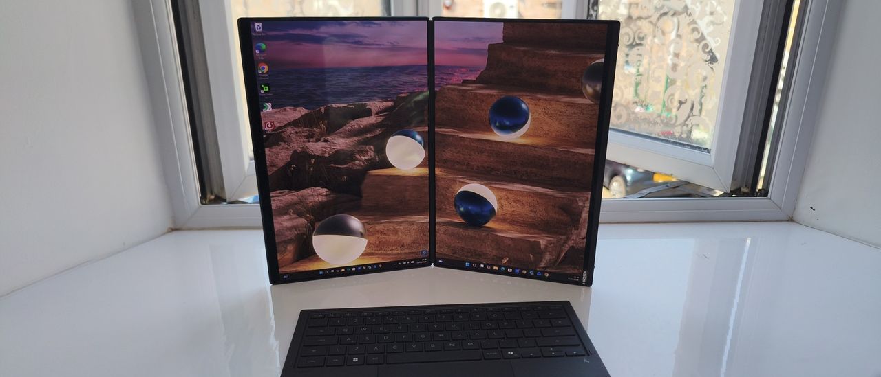 The ASUS Zenbook Duo standing vertically like an open book, displaying a continuous ocean sunset wallpaper split across two portrait screens, with the detached wireless keyboard resting in front.