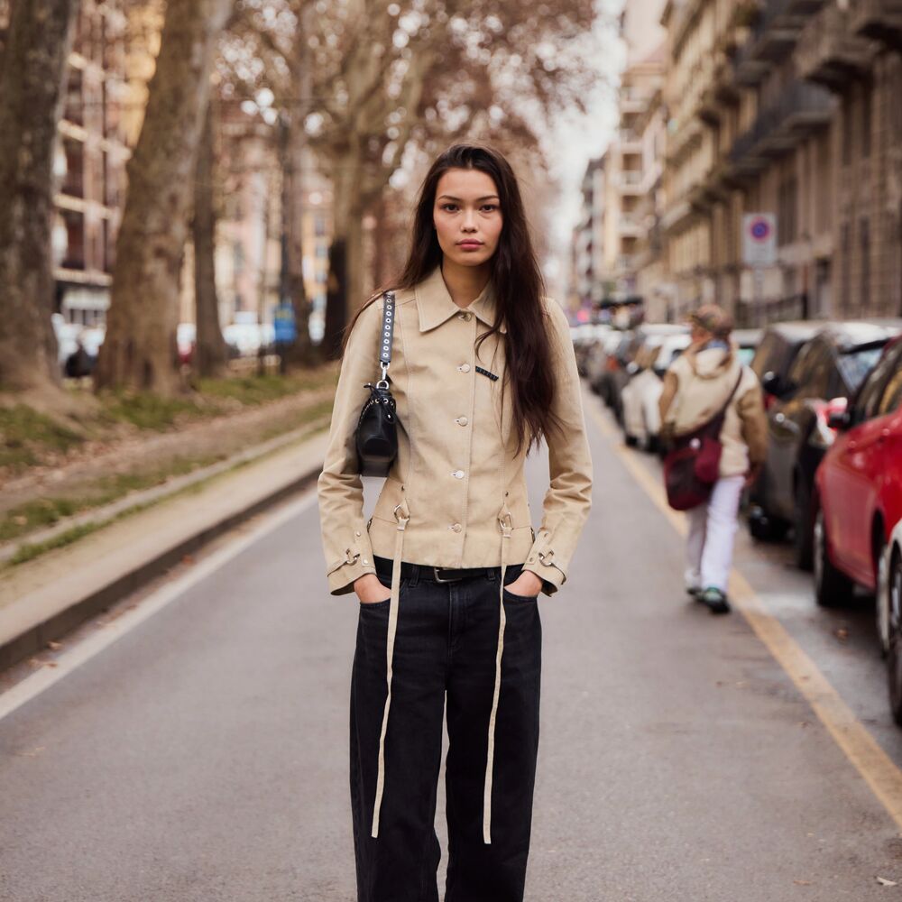 Woman wears cropped fitted trench style coat and black trousers while posing in the street. 