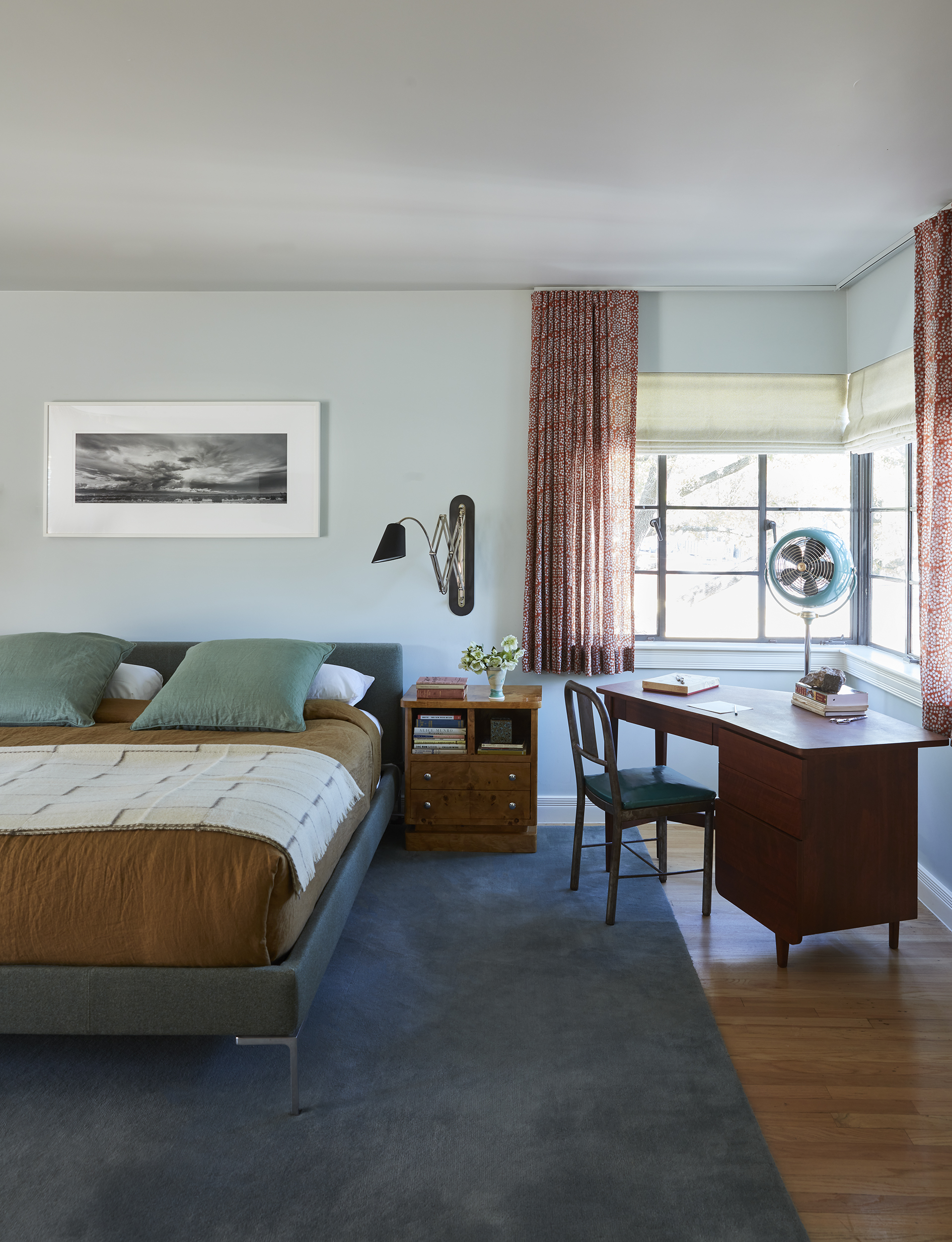 Image of a large, light blue bedroom with a bed in the middle and a wooden desk in the corner. The bed is blue with a brown duvet and teal pillows. There are windows above the desk that have short, red curtains.