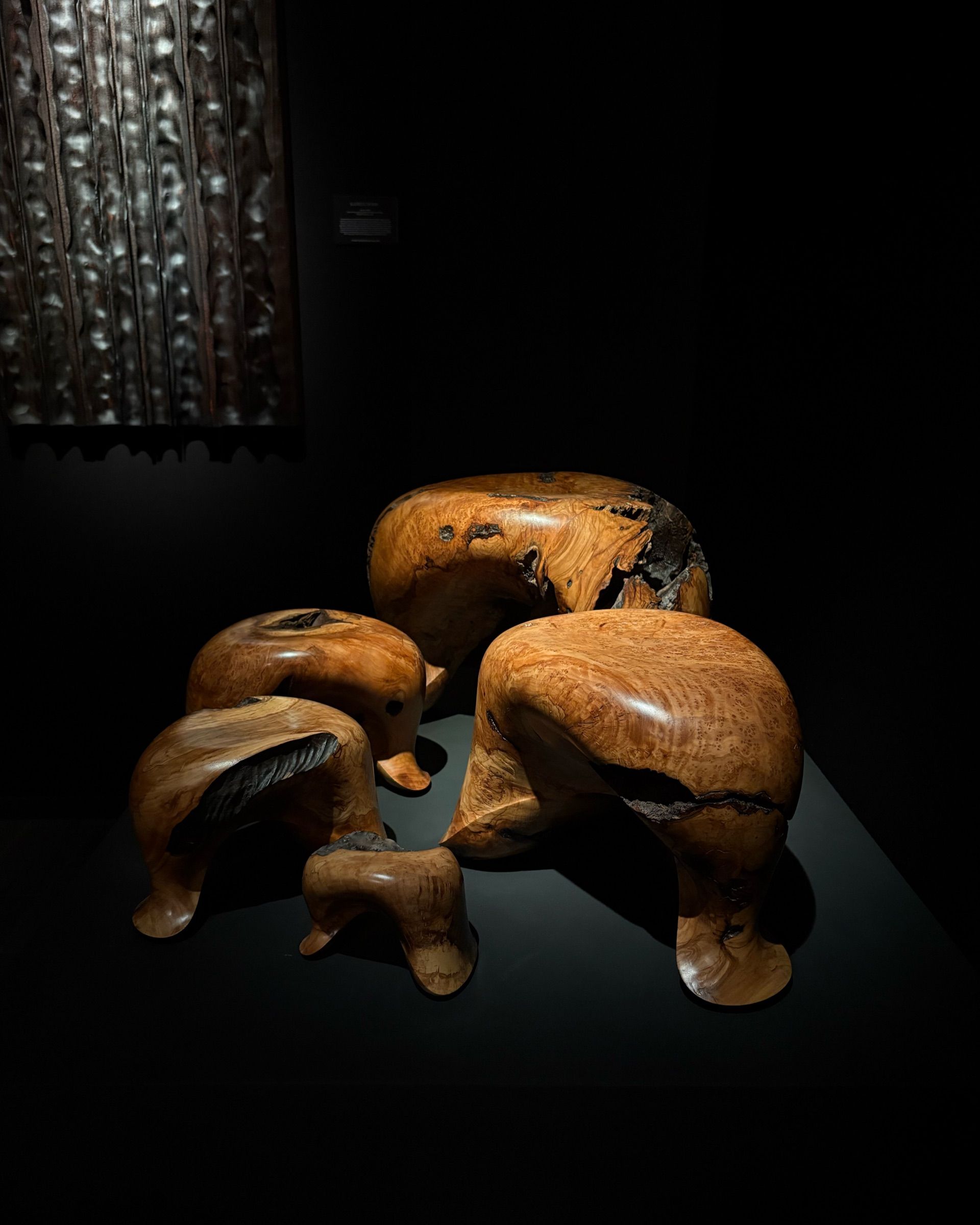 A series of legged, creaturesque wooden stools sit in a dark room illuminated by a spotlight.