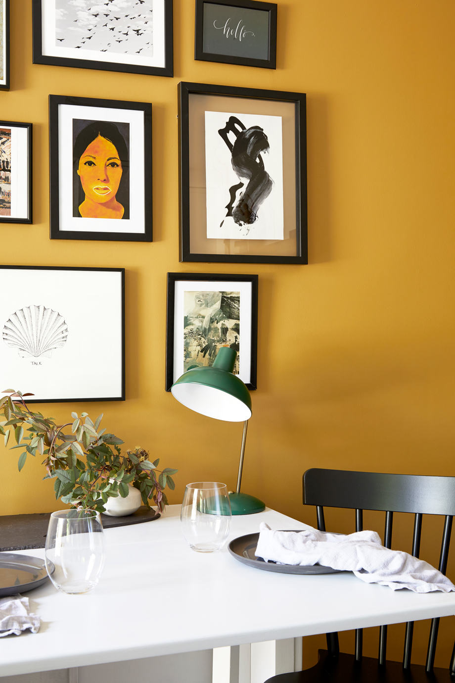a yellow dining room with artwork in a gallery wall and a green lamp on the dining table
