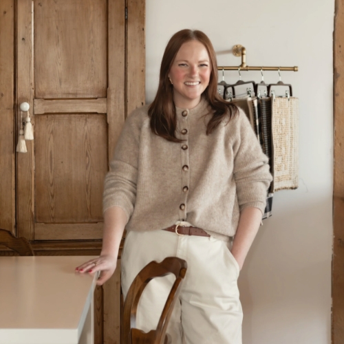 Image of a woman in a beige sweater and white pants standing in front of a wooden cupboard.