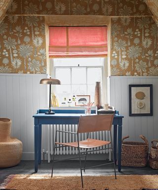 Blue desk with leather and metal chair, window with red blind, ochre floral wallpaper, light blue painted paneling, metallic table lamp, textured rug and woven baskets