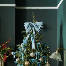 A Christmas tree in a dark green-painted dining room decorated with blue velvet ribbons and a large patterned blue bow used as a tree topper