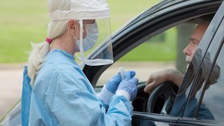 Dressed in full protective gear, a healthcare worker administers a COVID-19 test to a person in their vehicle. 