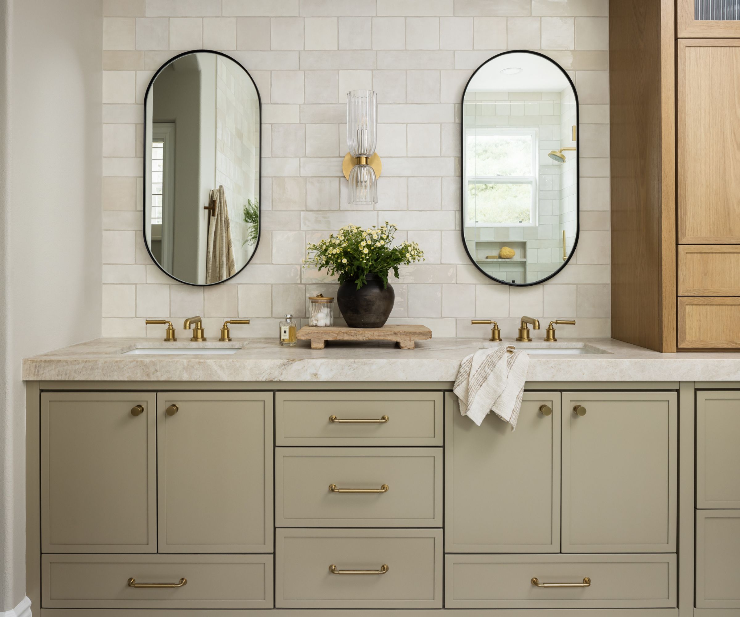 A green bathroom vanity with marble counters, a tiled backsplash, the black pill-shaped mirrors, and one wall light between them