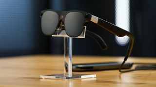 A photo of the XREAL Project Aura glasses sitting on a plastic pedestal, with dark lenses and a cord connected to a computing device with trackpad.