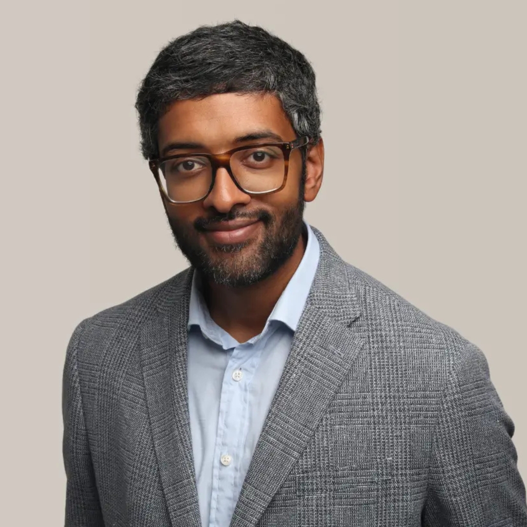 Image of Dr Sreedhar Krishna smiling with short black hair and beard, wearing glasses, grey check blazer and light blue shirt