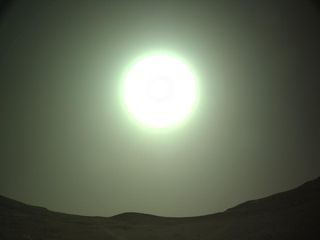 a bright, green-hued fireball of white hangs in the hazy sky above a low skyline of rolling hills.